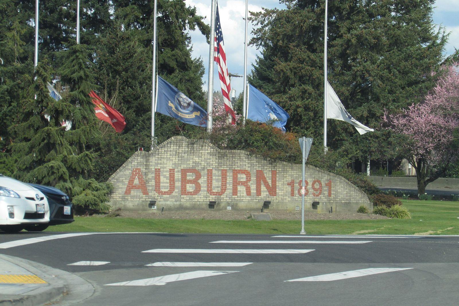 Brick Auburn 1891 welcome sign with flags and evergreen trees behind it